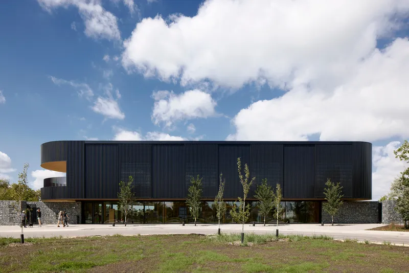 Front elevation showing the curved black metal upper volume cantilevered over a gabion stone base with young trees