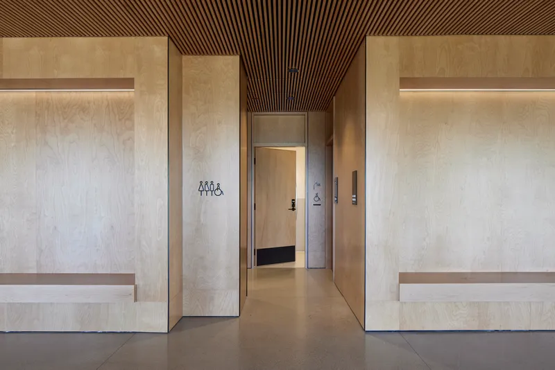 Corridor with light plywood wall panels and integrated benches beneath a slatted wood ceiling
