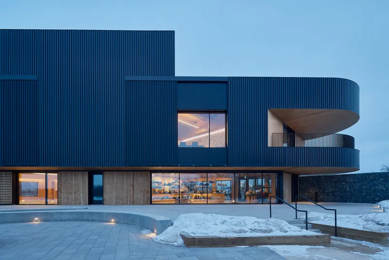 Ribbed blue metal facade with cantilevered upper volume illuminated at dusk in winter snow