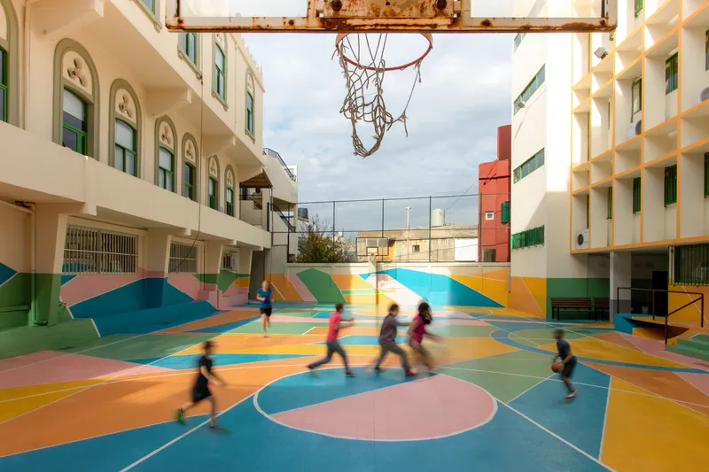 Basketball hoop suspended above the courtyard as children in motion play across the painted surface