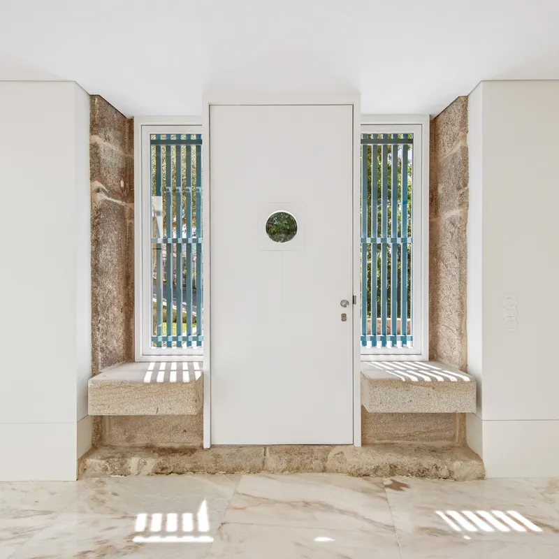 White door with circular window framed by weathered stone reveals and vertical slatted sidelights casting shadow stripes