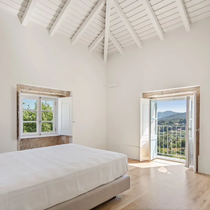 Bedroom with pitched white ceiling and two stone-framed openings overlooking hillside landscape in daylight