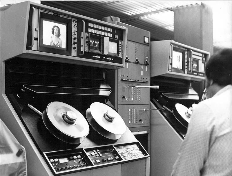 Operator working at a console with reel-to-reel tape drives and monitor screens in a ribbed ceiling room