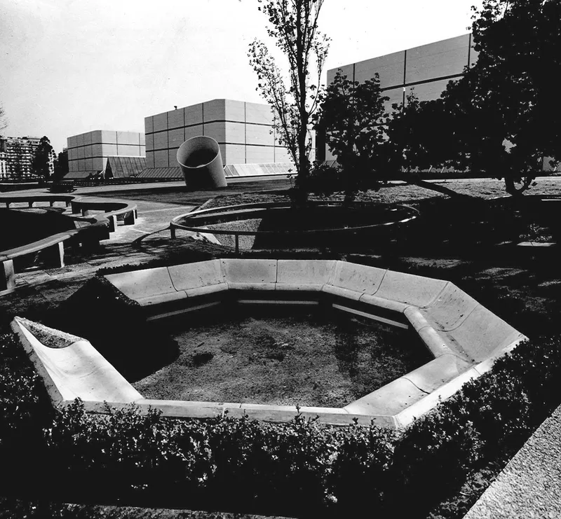 Octagonal concrete seating area with planted ground cover in a landscaped plaza with gridded facade beyond