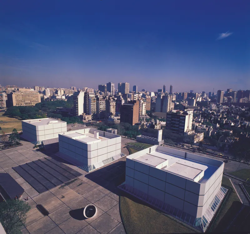 Aerial view of white paneled volumes with cylindrical sculpture on paved plaza overlooking dense urban skyline