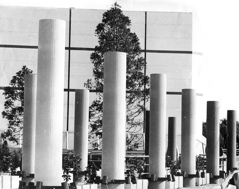 Grid of white cylindrical columns with horizontal bands and trees visible beyond