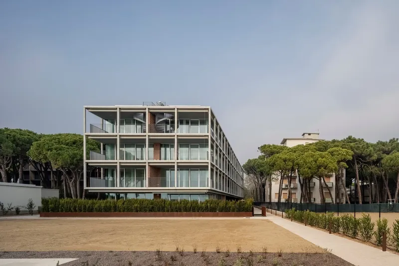 Oblique view across the sand courtyard showing the building flanked by dense pine groves under grey sky