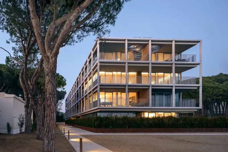 Facade with illuminated balconies and vertical timber screens viewed from pathway flanked by pine trees at dusk