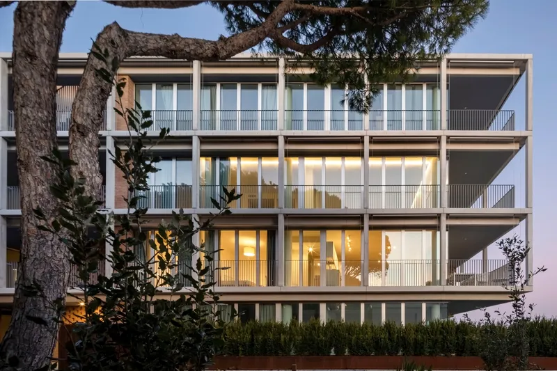 Four-story facade section with glazed walls and concrete frame seen through leaning pine tree trunks at twilight