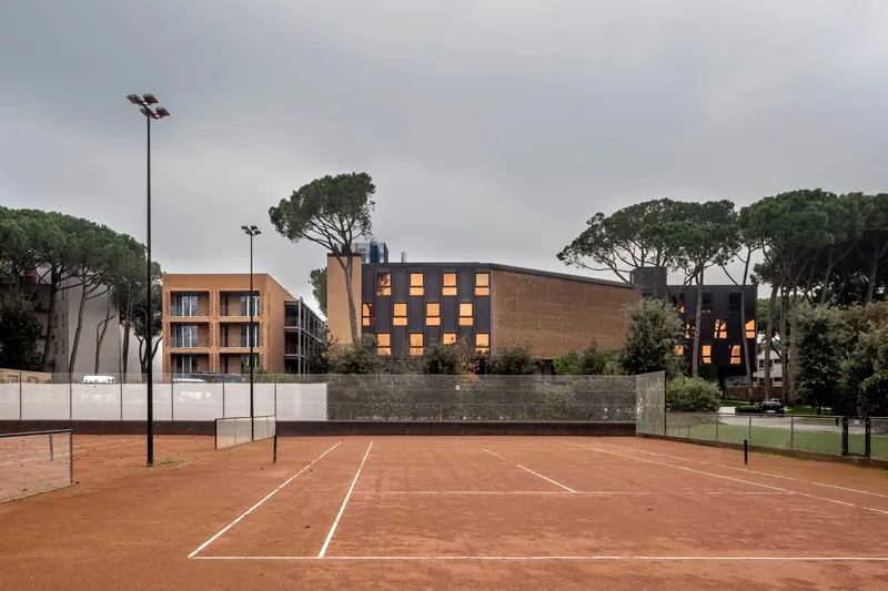 Perforated brick facade with illuminated windows viewed across a red clay tennis court at dusk