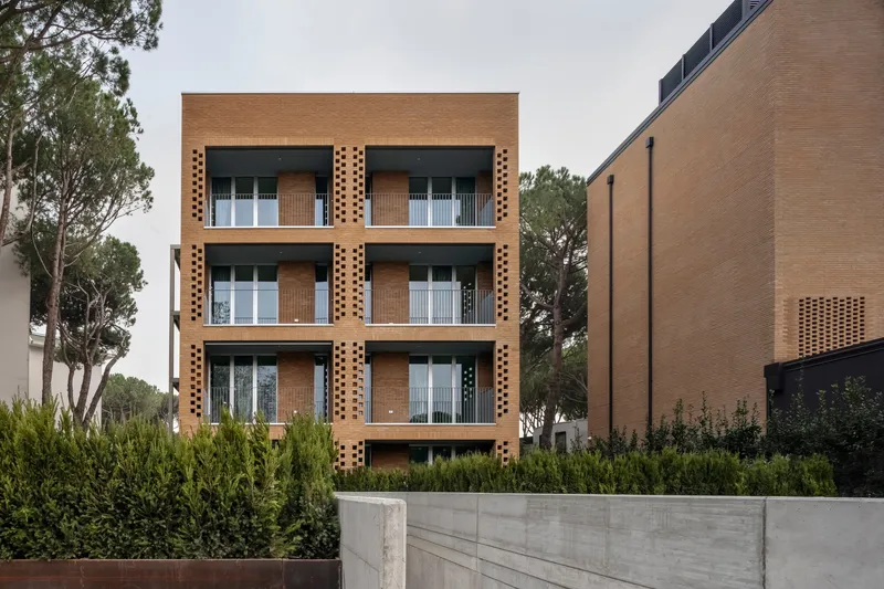 Four-story perforated brick facade with recessed balconies behind a concrete retaining wall and planted hedge