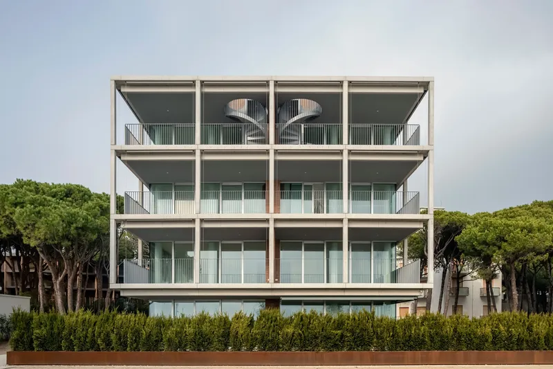 Frontal view of the four-storey concrete frame facade with open balconies behind a hedge of green shrubs