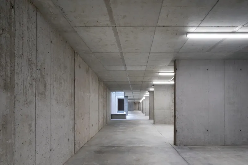 Board-formed concrete corridor with exposed ceiling beams and fluorescent light strips leading to a distant opening