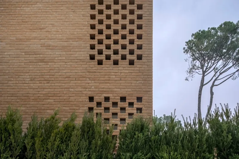 Buff brick facade with chequered pattern of void openings beside pine trees under overcast sky