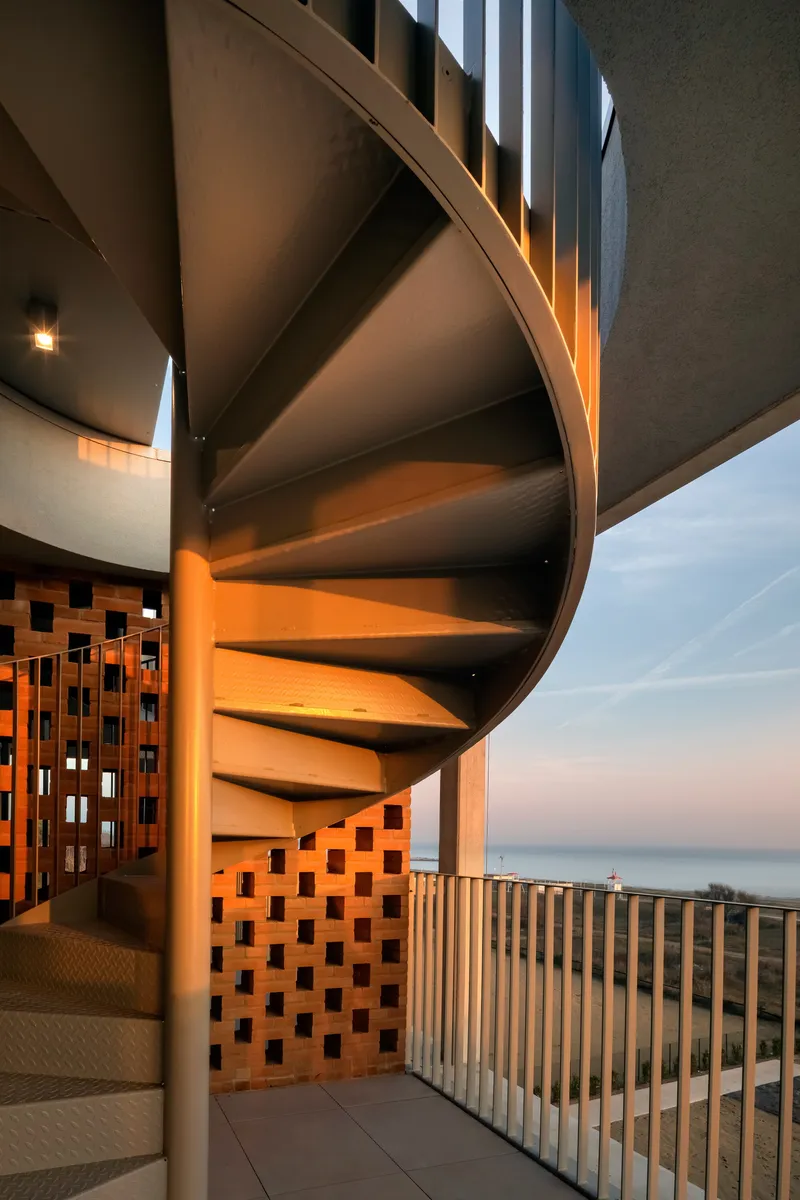 Spiral stair rising through vertical slat screen with perforated brick wall illuminated at twilight