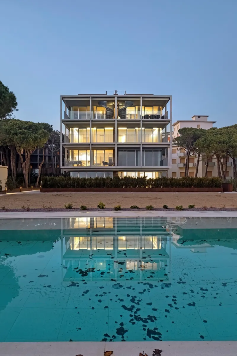 Four-story glass and steel facade reflected in the pool at twilight with pine trees flanking