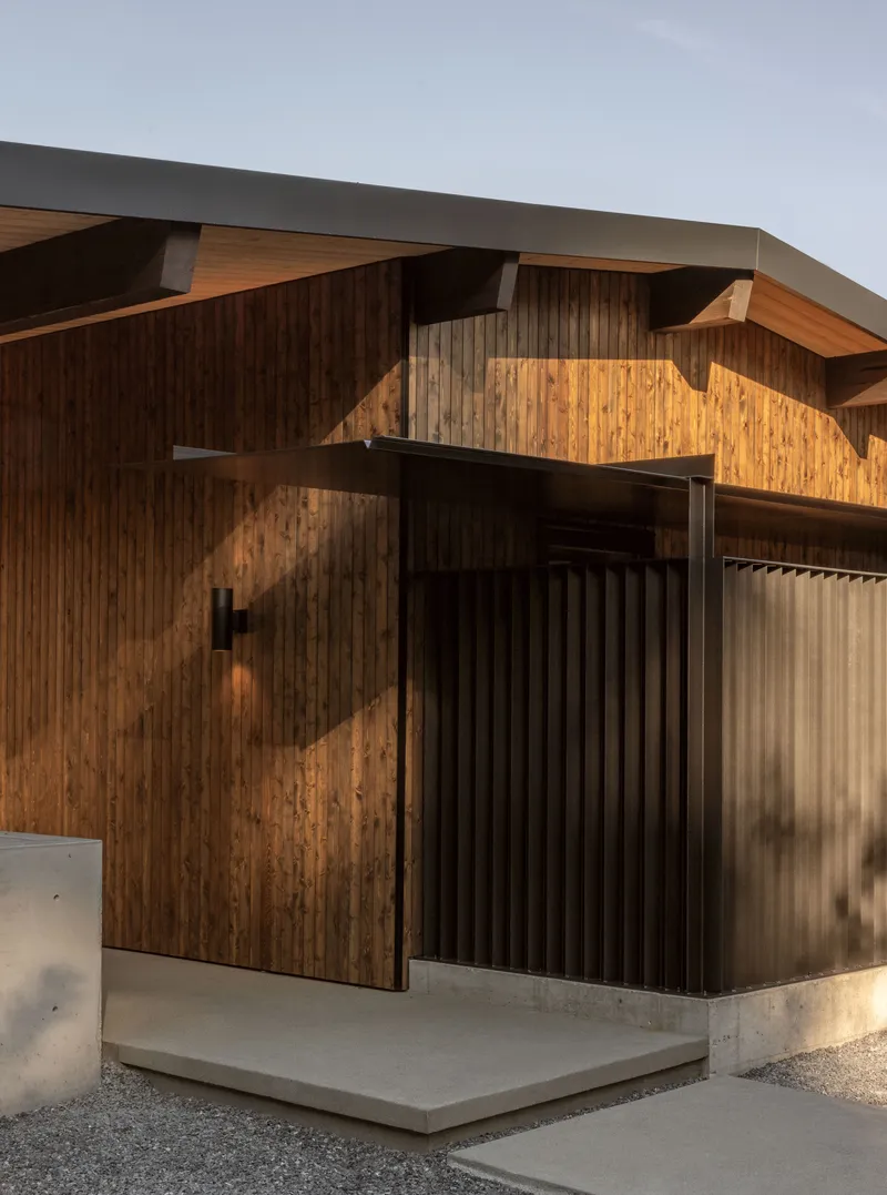 Entry elevation showing vertical timber siding with diagonal sunlight patterns and concrete plinth below