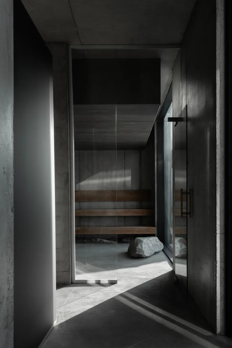 Glass door entry to a timber sauna with a boulder on concrete floor illuminated by vertical light slot