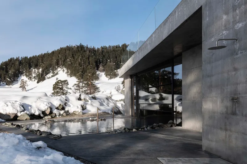 Concrete pavilion with glass walls and reflecting pool beside a snow-covered hillside with evergreens