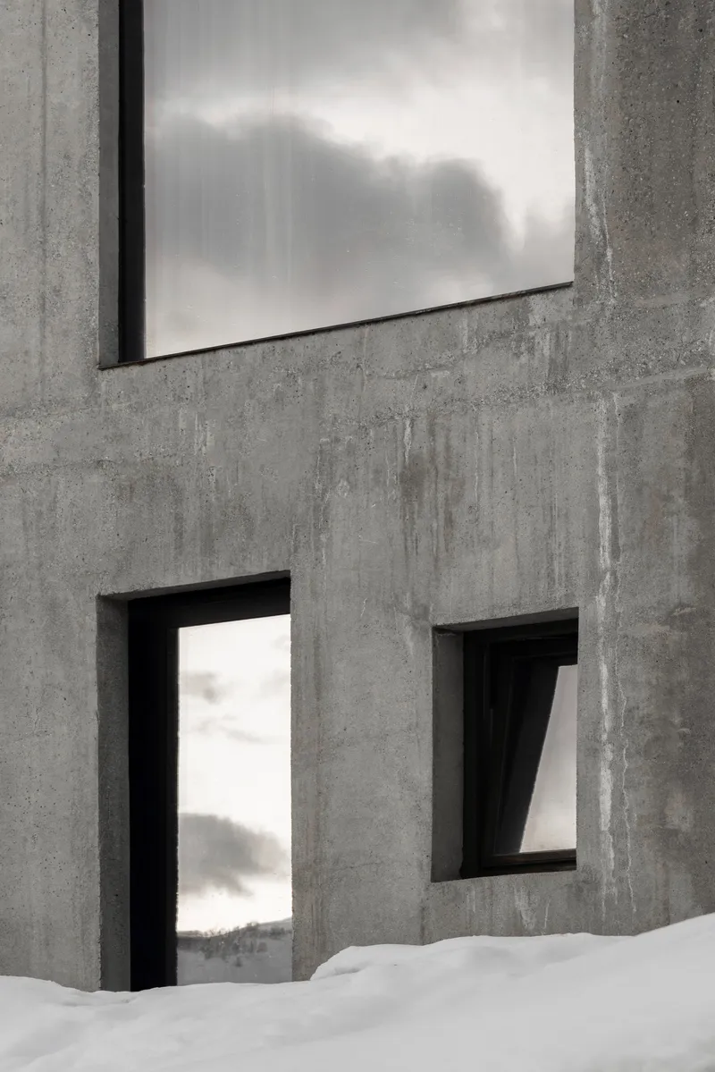 Board-formed concrete facade with rectangular window openings and deep reveals beside snowdrift
