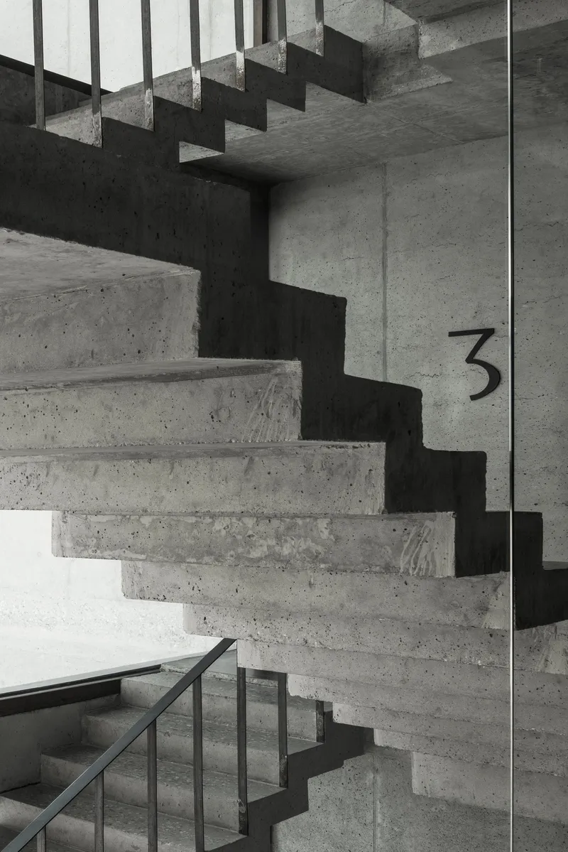 Folded concrete staircase with exposed formwork texture and steel handrail under natural light from a skylight