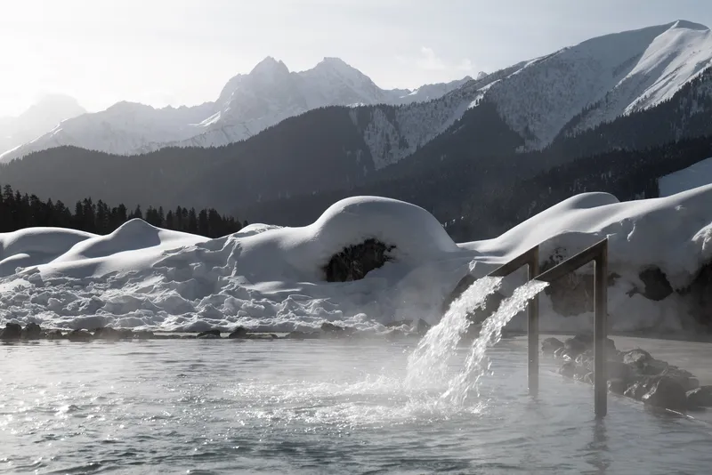 Outdoor pool with cascading water feature set against snow-covered landscape and mountain peaks at dusk