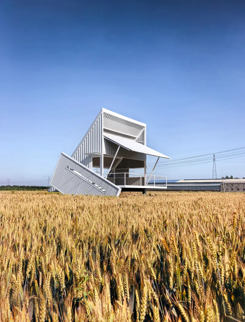 White corrugated metal structure with tilted roof planes rising above a harvested wheat field
