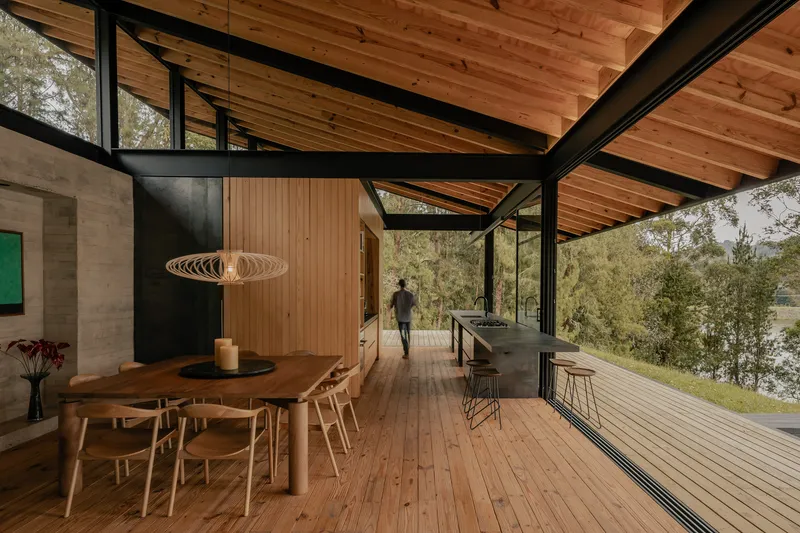 Open-plan dining and kitchen space with timber ceiling, black steel structure, and figure walking toward the landscape