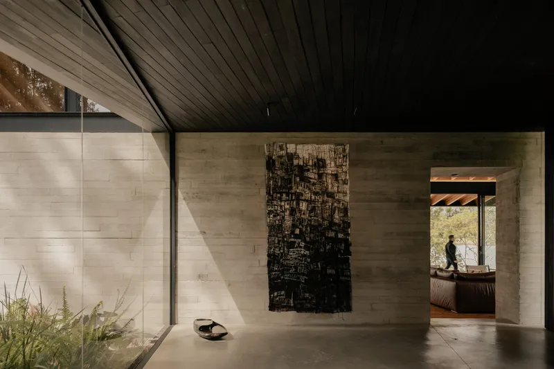 Gallery hallway with board-formed concrete walls, dark timber ceiling, and figure silhouetted in distant doorway