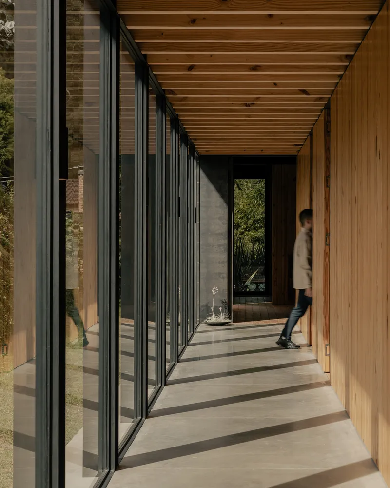 Glazed corridor with person walking on concrete floor beneath exposed timber joists casting shadows