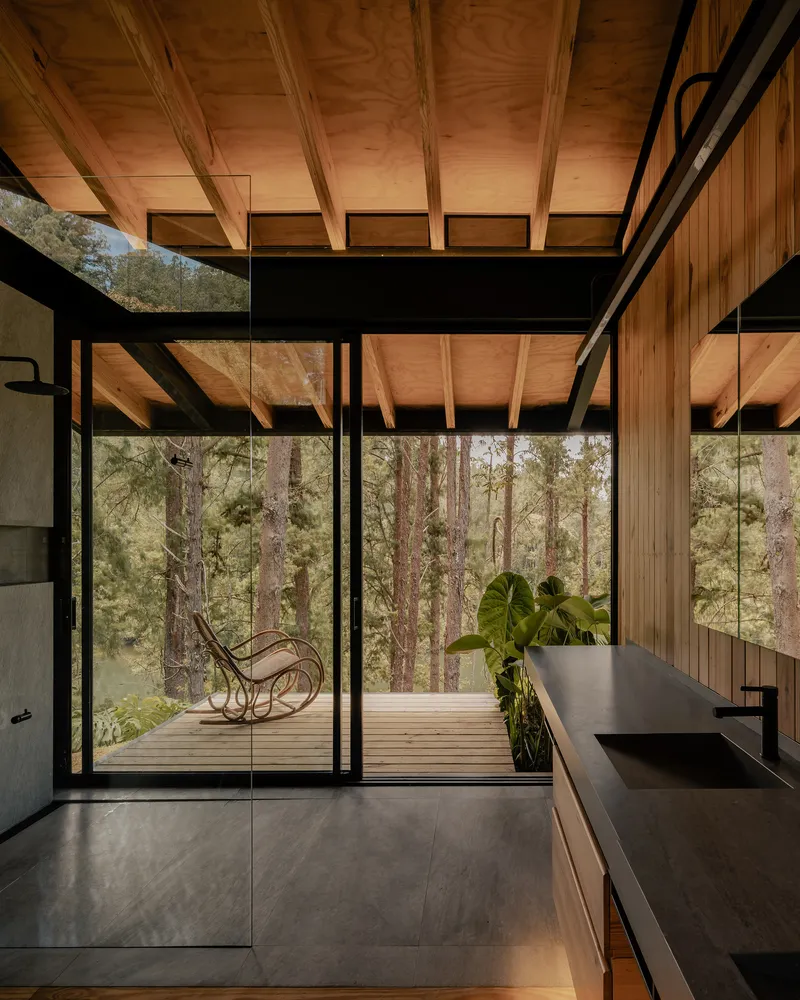 Interior view through floor-to-ceiling glass walls opening to a timber deck with rocking chair among trees