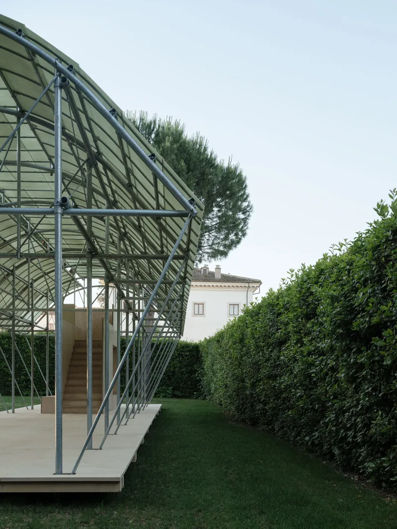 Close-up of the exposed triangulated steel frame supporting the translucent panel roof against hedges