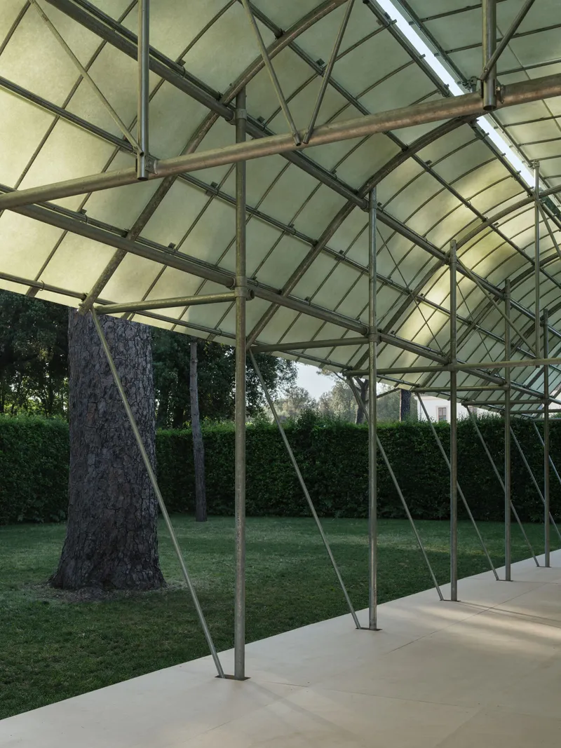 Covered pavilion with translucent fabric roof and steel frame supported by diagonal columns in a grassy clearing
