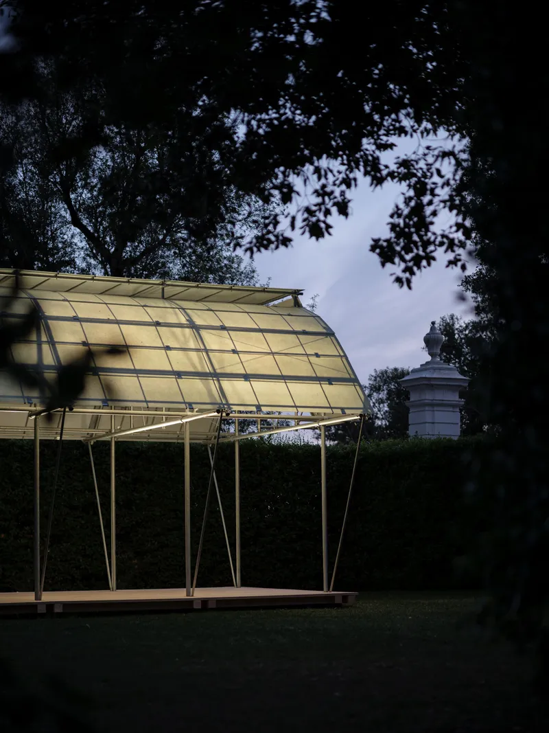Curved translucent roof structure supported by angled steel legs beneath overhanging trees at dusk