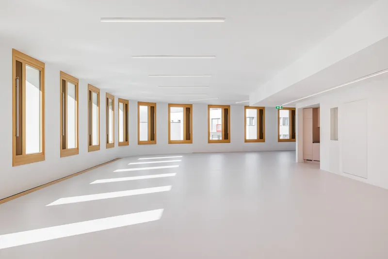 Empty white interior with timber-framed windows casting sunlight across the pale floor