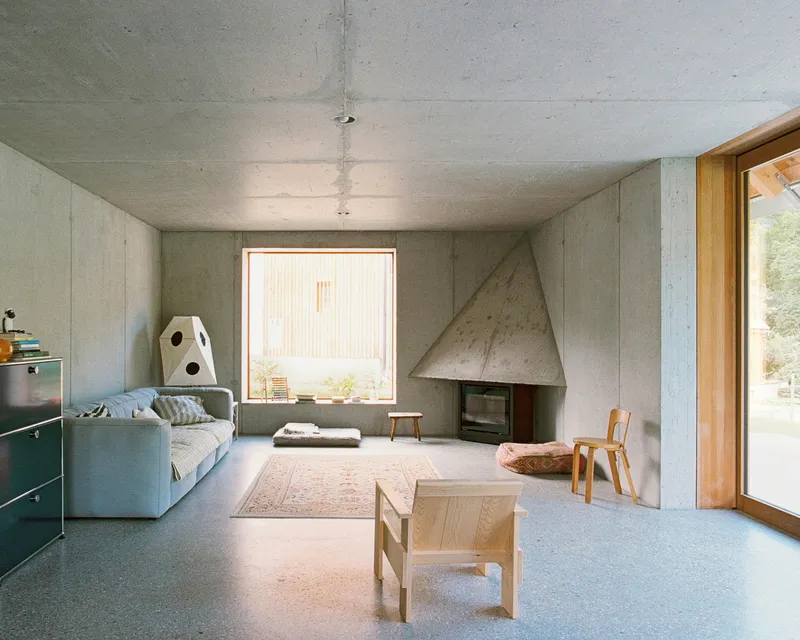 Living room interior with exposed concrete walls, geometric fireplace, and single square window framing the landscape