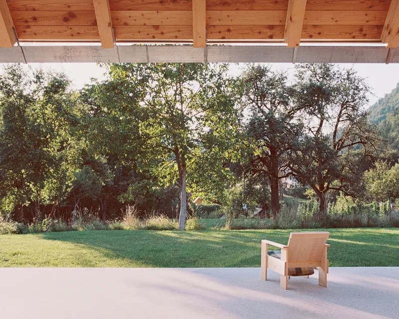 Covered terrace with exposed timber beams and single wood lounge chair facing a lawn and orchard