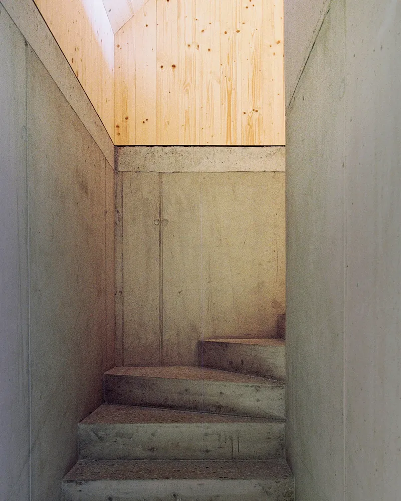 Narrow concrete stairwell with board-formed walls leading upward to a skylit plywood ceiling