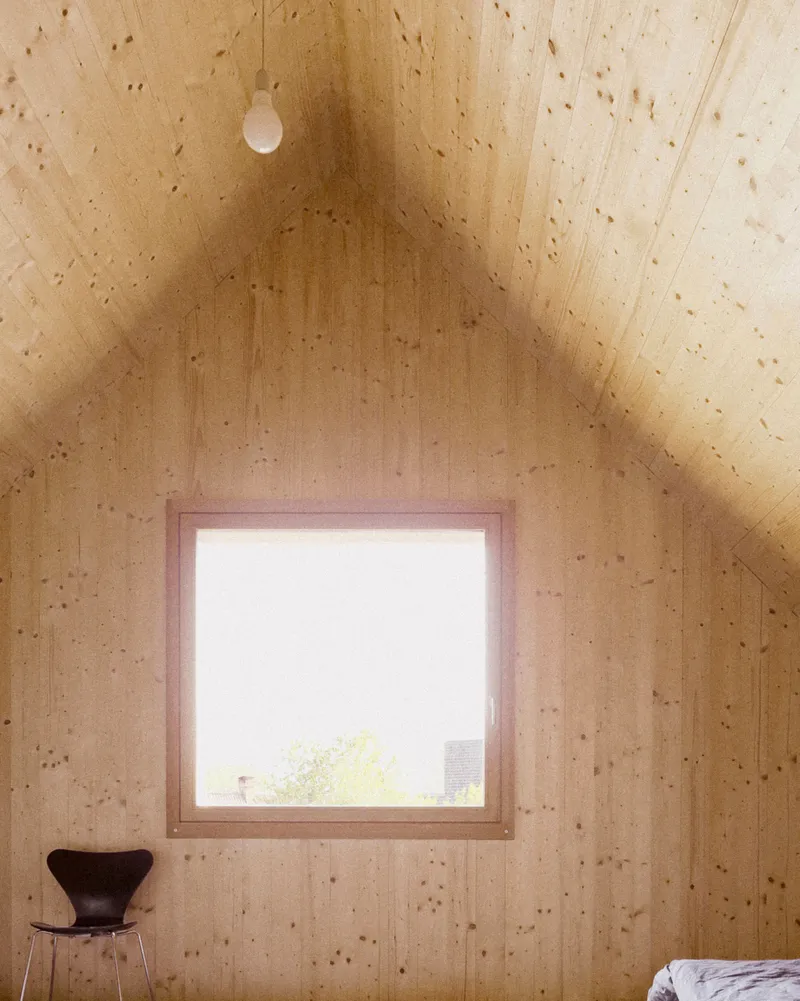 Gabled bedroom interior with plywood walls and a square window framing trees outside