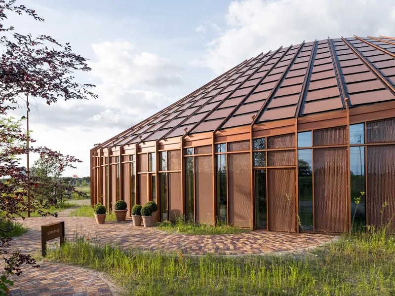Curved facade with copper-toned metal panels and vertical glazing beside a brick paved path with potted shrubs