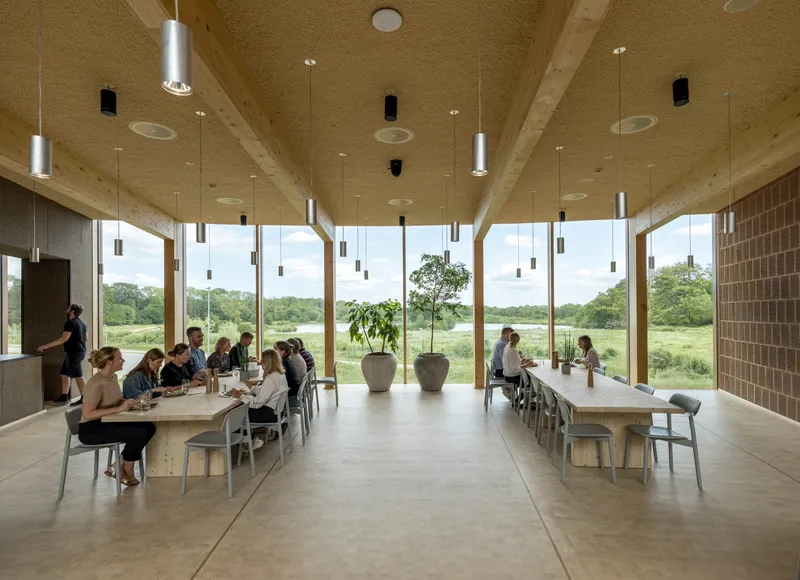 Interior dining space with long timber tables and floor-to-ceiling glazing framing a view of open meadow