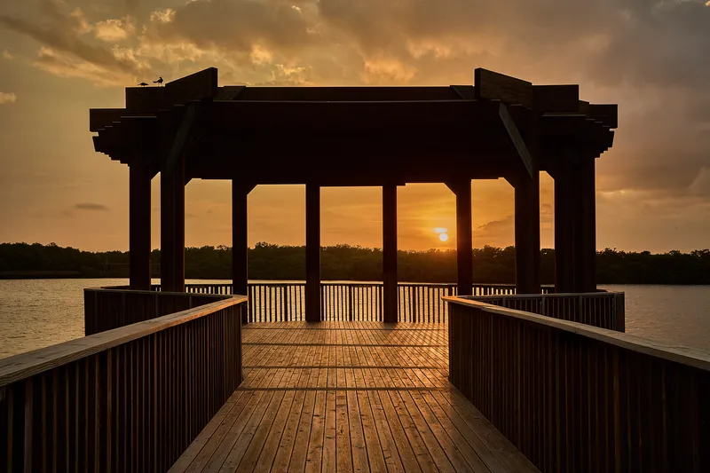 Timber deck pavilion with vertical slat railings and post-and-beam roof silhouetted at sunset over water
