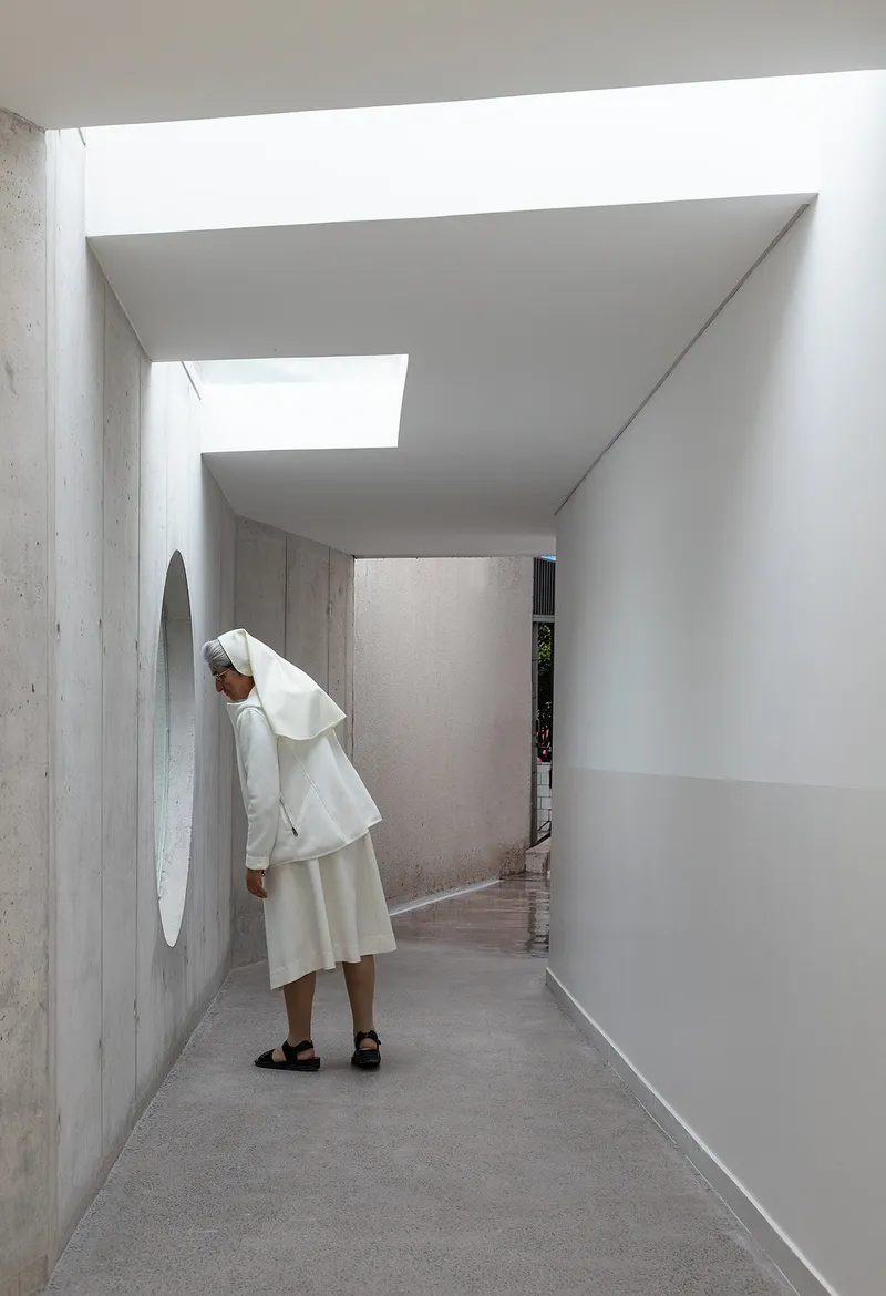 Skylit corridor with exposed concrete walls and a person standing beneath layered ceiling planes