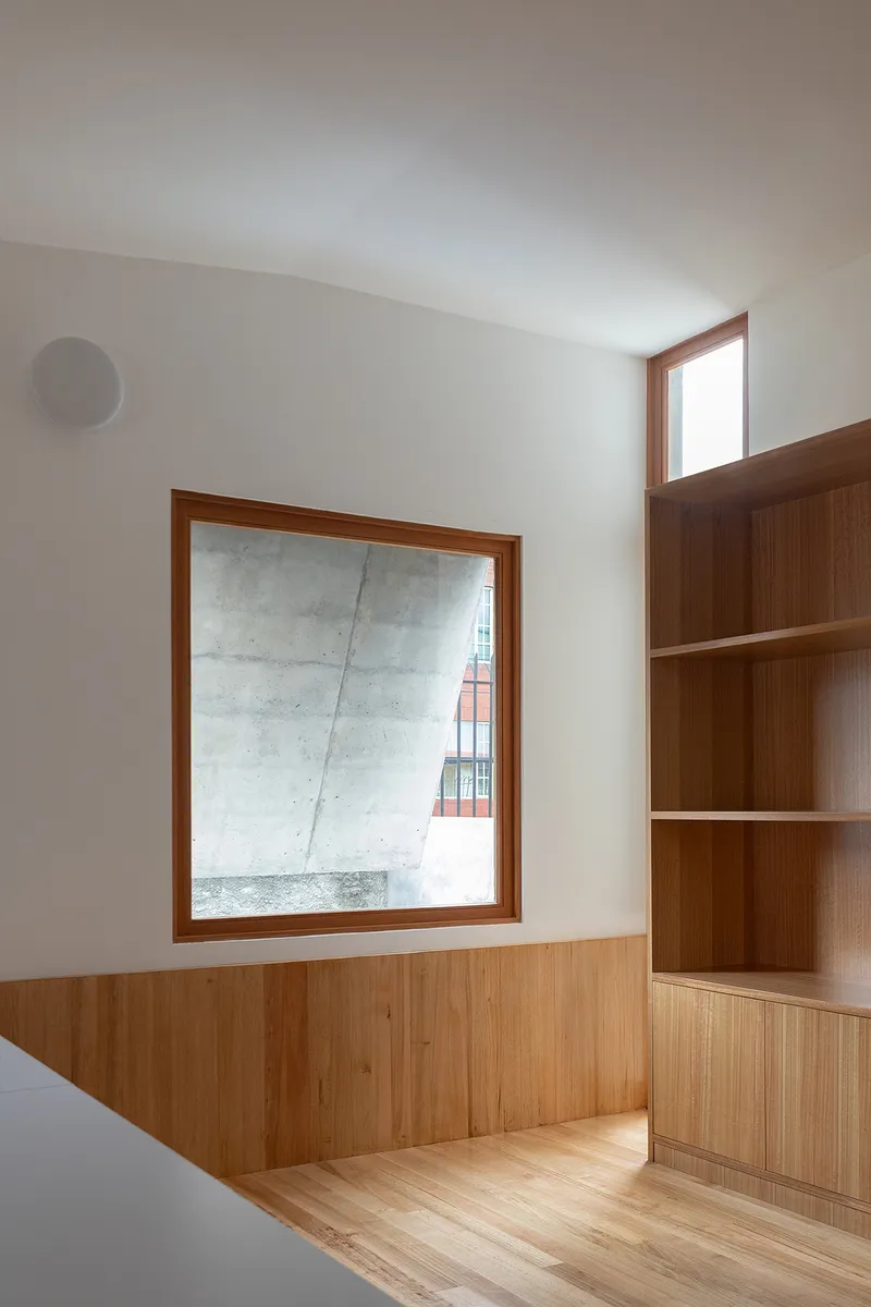 Timber-paneled room with built-in shelving and a square window overlooking an adjacent concrete wall