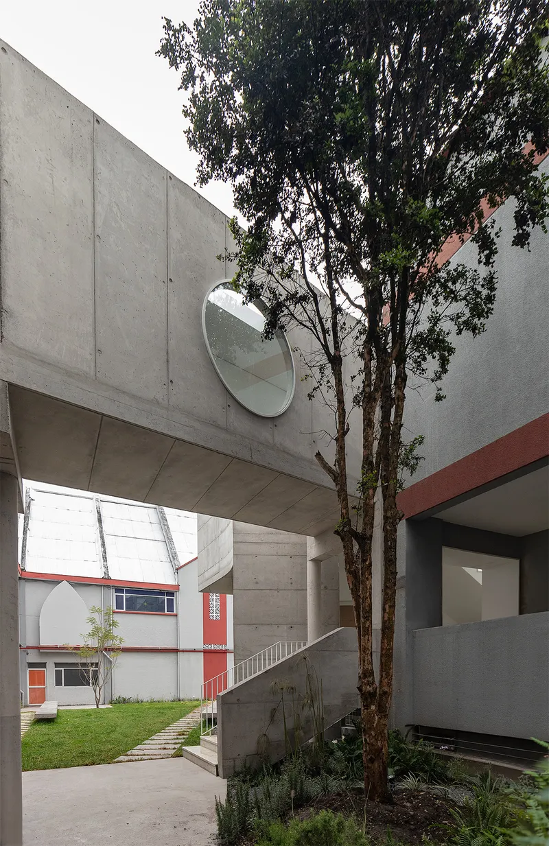 Courtyard view with concrete bridge overhead, circular mirror on the wall, and tree growing between volumes