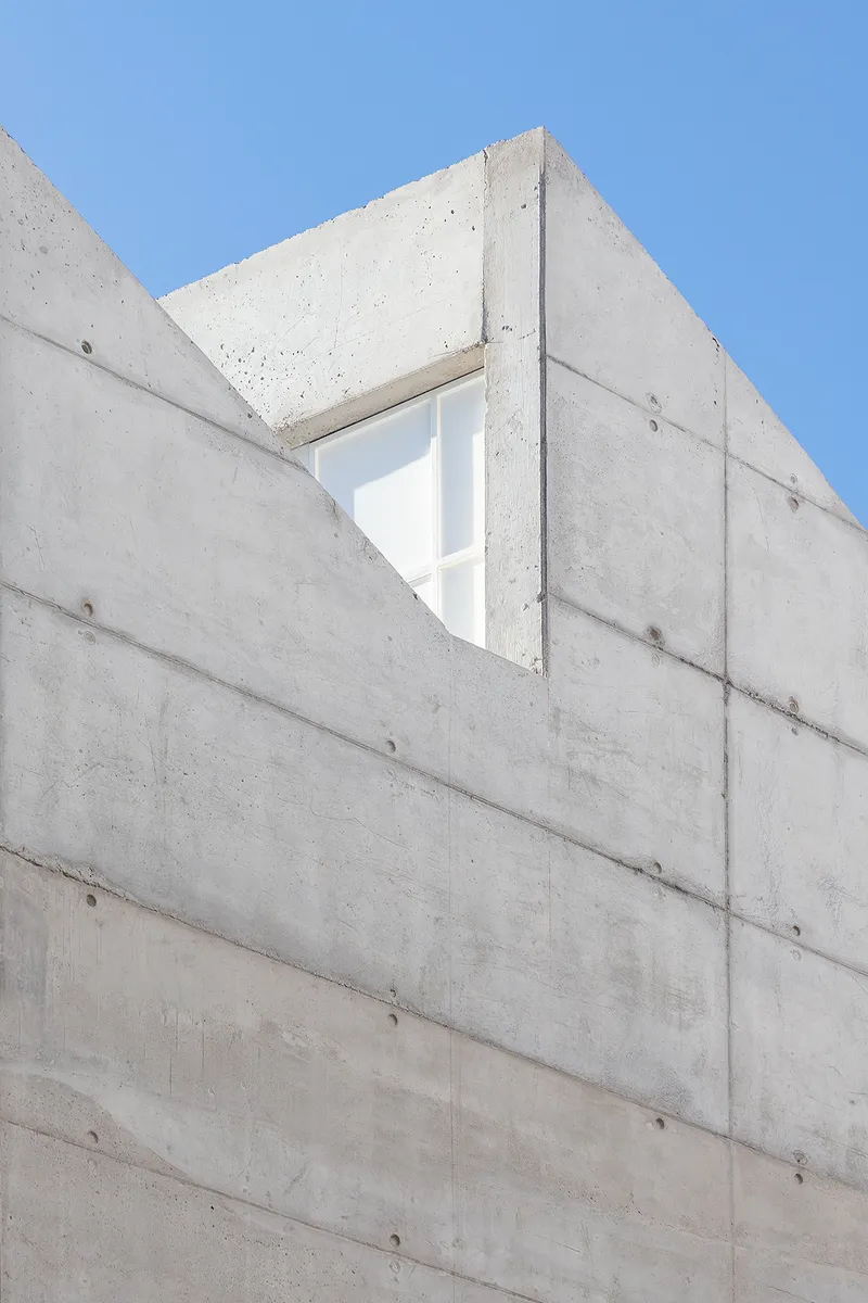 Board-formed concrete wall with triangular window opening under a clear blue sky