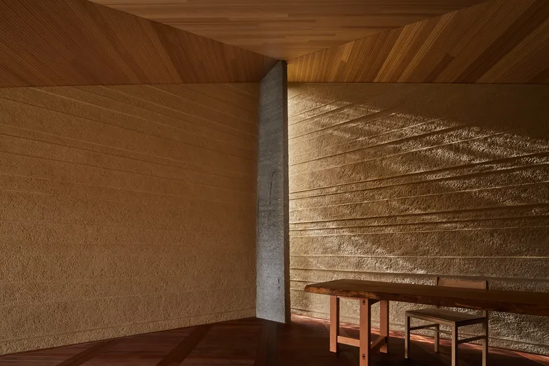 Timber seating below horizontally textured rammed earth wall with concrete column and wood ceiling