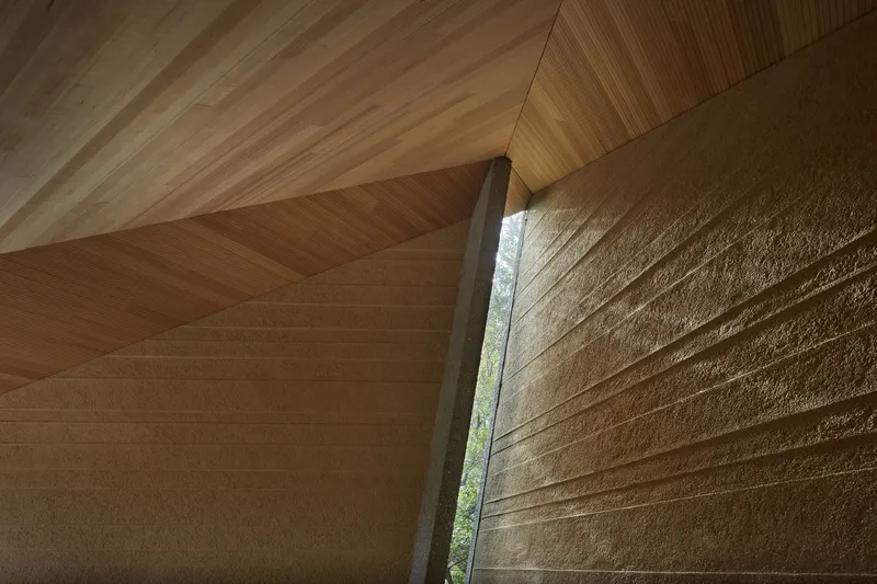 Angled timber ceiling meeting textured stone wall at a vertical window slit revealing foliage