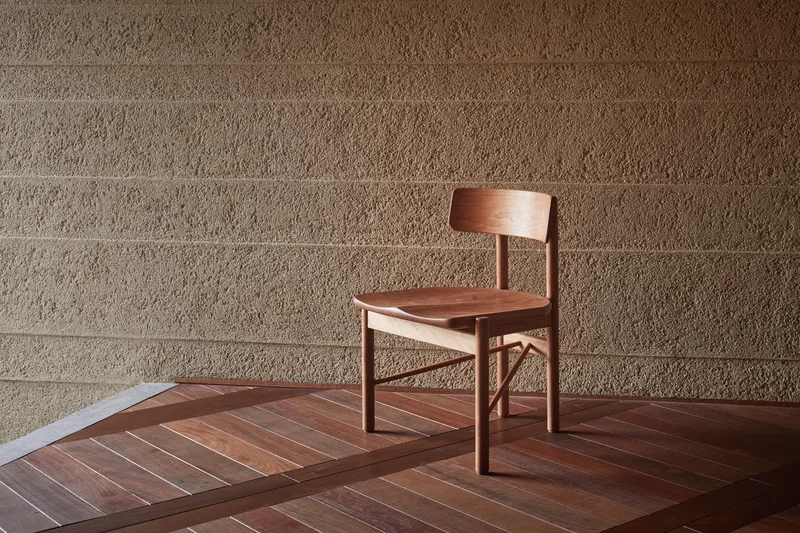 Timber chair on geometric wood floor against horizontal-grooved stone block wall in afternoon light
