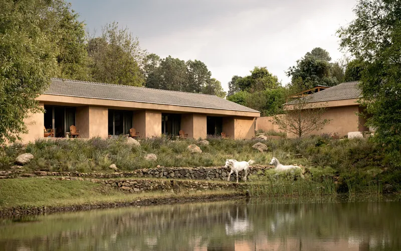 Rammed earth pavilions beside a pond with white goats grazing on the sloped bank